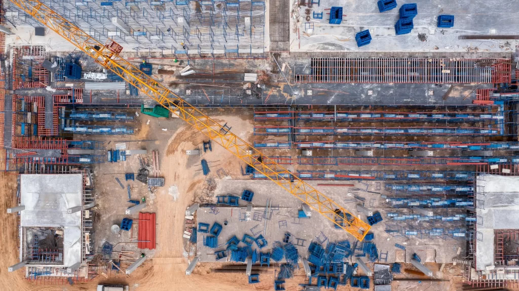 Aerial view of a construction site featuring various machinery, materials, and organized stacks of containers. The layout shows ongoing development with a mix of dirt and concrete surfaces.