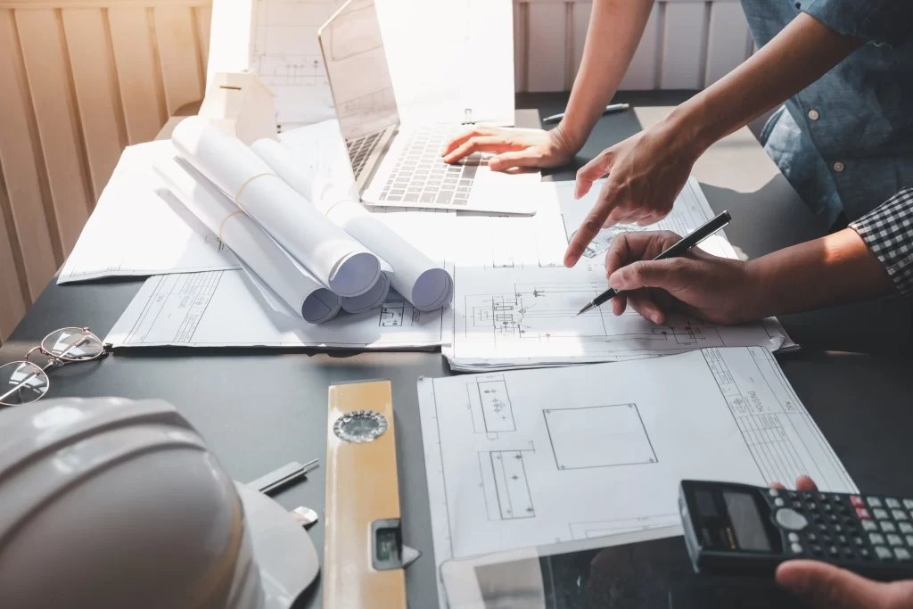 Two hands point at architectural plans spread across a table, accompanied by a laptop and a calculator. A hard hat and a level are also visible, indicating a construction or design context.