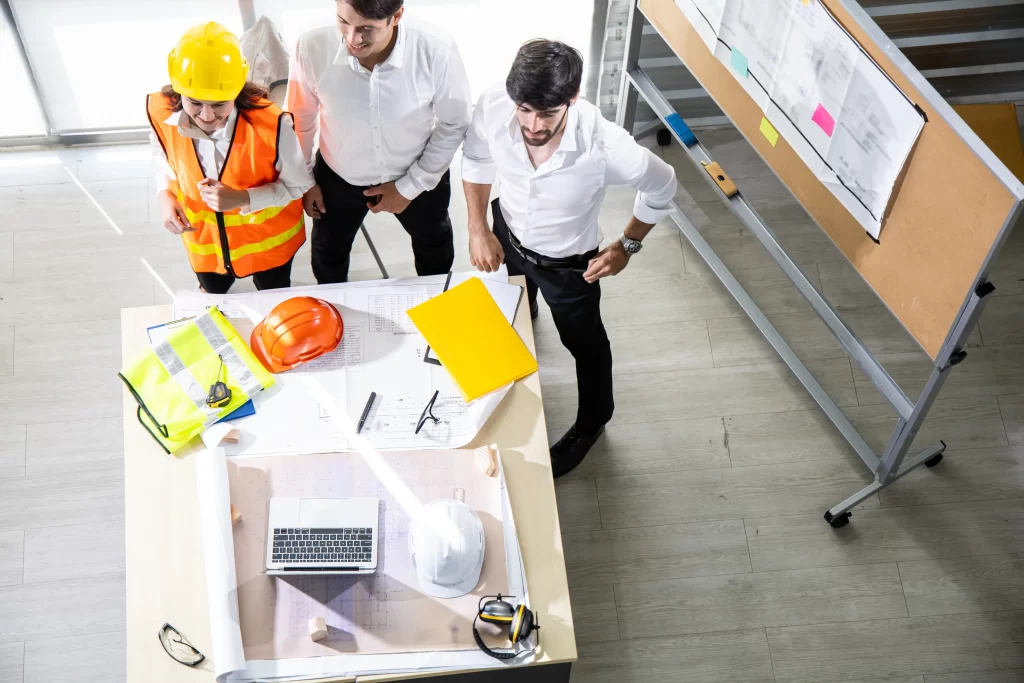 Three professionals, including a construction worker in a safety vest and helmet, discuss plans and materials on a table in a bright office space. Various construction tools and documents are visible around them.