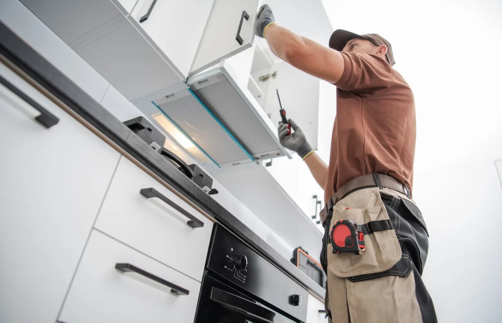 A worker installs a kitchen cabinet, using tools and wearing gloves, in a modern kitchen setting. The image captures the process of home improvement.