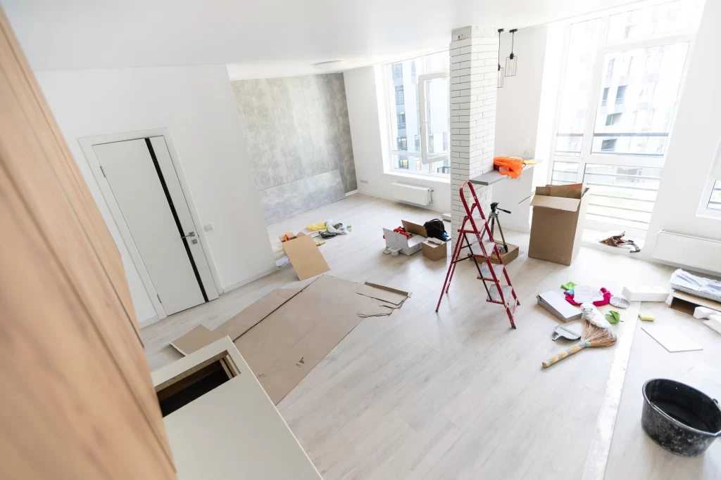 A spacious room undergoing renovation, featuring scattered tools, materials, and a ladder, with large windows allowing natural light.