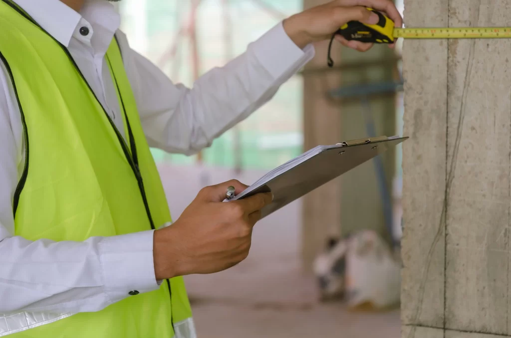 A construction worker in a safety vest measures a column with a tape measure while holding a clipboard. The background shows an under-construction site.