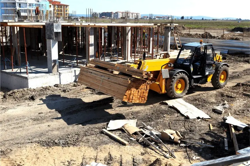 A construction vehicle is lifting materials at a building site, with a partially constructed structure in the background. The ground is uneven and scattered with construction debris.
